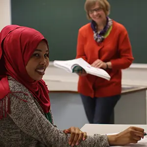 Opiskelija hymyilee ja kouluttaja taustalla hymyilee kirja kädessään.